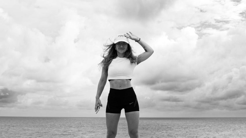 Woman in sportswear stands confidently by the sea against a cloudy sky.